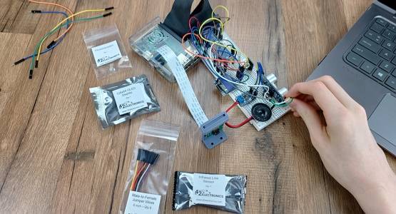 Hand working on a breadboard circuit featuring a Raspberry Pi camera, speaker, audio amplifier, OLED screen, ultrasonic sensor, GPIO header, and a variety of jumper wires; packaged electronic components and laptop are also on the table