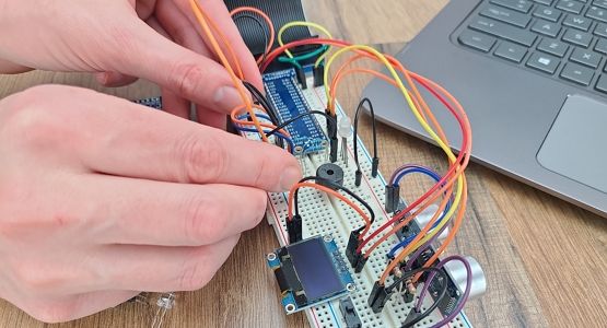 On wood table two hands work on a breadboard circuit next to a laptop; circuit includes GPIO header and ribbon, piezo speaker, RGB LED, OLED screen, slide switch, ultrasonic sensor, and jumperwires; laptop, phototransistor, and temperature sensor sit next to circuit