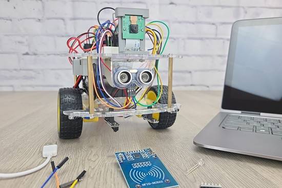On grey wood table with white brick background sits a robot alongside a laptop, USB audio connector, jumper wires, temperature sensor, phototransistor, and RFID reader