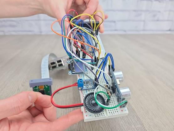 Hands holding up a breadboard circuit featuring a Raspberry Pi camera, speaker, audio amplifier, OLED screen, ultrasonic sensor, GPIO header, and a variety of jumper wires