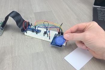 Hand holding RFID tag next to RFID reader on breadboard circuit demonstrating how it is used; RFID card and laptop on table next to circuit