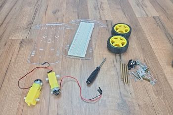 Robot chassis parts sitting on wood table; parts include upper clear plastic chassis with breadboard; lower clear plastic chassis; two DC motors, one screwdriver; two wheels, one rear caster with ball, and assorted metal hardware