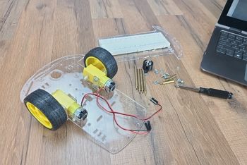 Lower clear plastic chassis sits on table next to laptop with two DC motors and two wheels installed; upper chassis with breadboard, screwdriver, rear caster, and assorted hardware sit on table