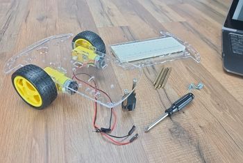 Sitting on wood table is a laptop, an upper chassis plate with breadboard, assorted hardware, and screwdriver; the motors, wheels and rear castor have been installed on the lower chassis plate