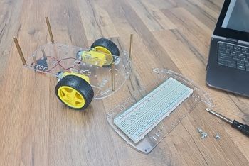 Partially constructed robot chassis sitting on wood table; includes lower chassis plaste, wheels, DC motors, rear caster, and metal standoffs; on table next to it sits upper chassis plate with breadboard, assorted hardware, screwdriver, and laptop