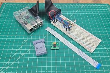 Breadboard circuit with basic electronic components and GPIO header installed sitting on green engineering mat; Raspberry Pi attached via GPIO cable; camera case, Raspberry Pi camera, and camera cable sitting next to circuit