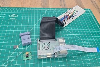Green engineering mat on table; sitting on mat is a Raspberry Pi with a camera cable installed; circuit with jumper wires in the background; uninstalled components on table including Raspberry Pi camera, camara case, and SD card; Raspberry Pi case is closed and camera cable has been threaded through opening in the case