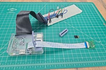 Green engineering mat on table; Raspberry Pi in clear case sits on mat; camera case has camera cable threaded through it and Raspberry Pi camera is attached to the end of the cable; breadboard circuit with basic components, GPIO ribbon and header, and jumper wires sits in the background