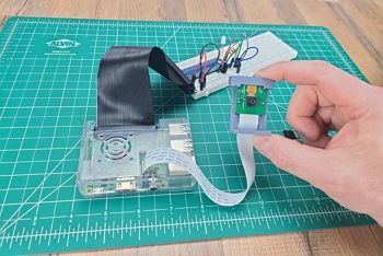 Green engineering mat on table ; hand holds up Raspberry Pi camera in case and attached to camera cable which is attached to a Raspberry Pi in a clear plastic case; the GPIO ribbon and header are attached to a breadboard circuit with basic comonents and jumper wires