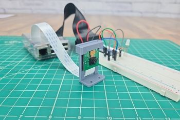 Green engineering mat on table; Raspberry Pi camera in case and attached to camera cable sits on mat; in background there is a Raspberry Pi, a GPIO ribbon and header, and a breadboard circuit with basic components and jumper wires