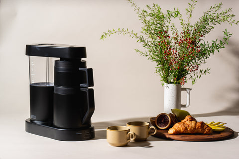 A black ratio six coffee brewer next to a pitcher with florals and a arrangement of snacks with cups.