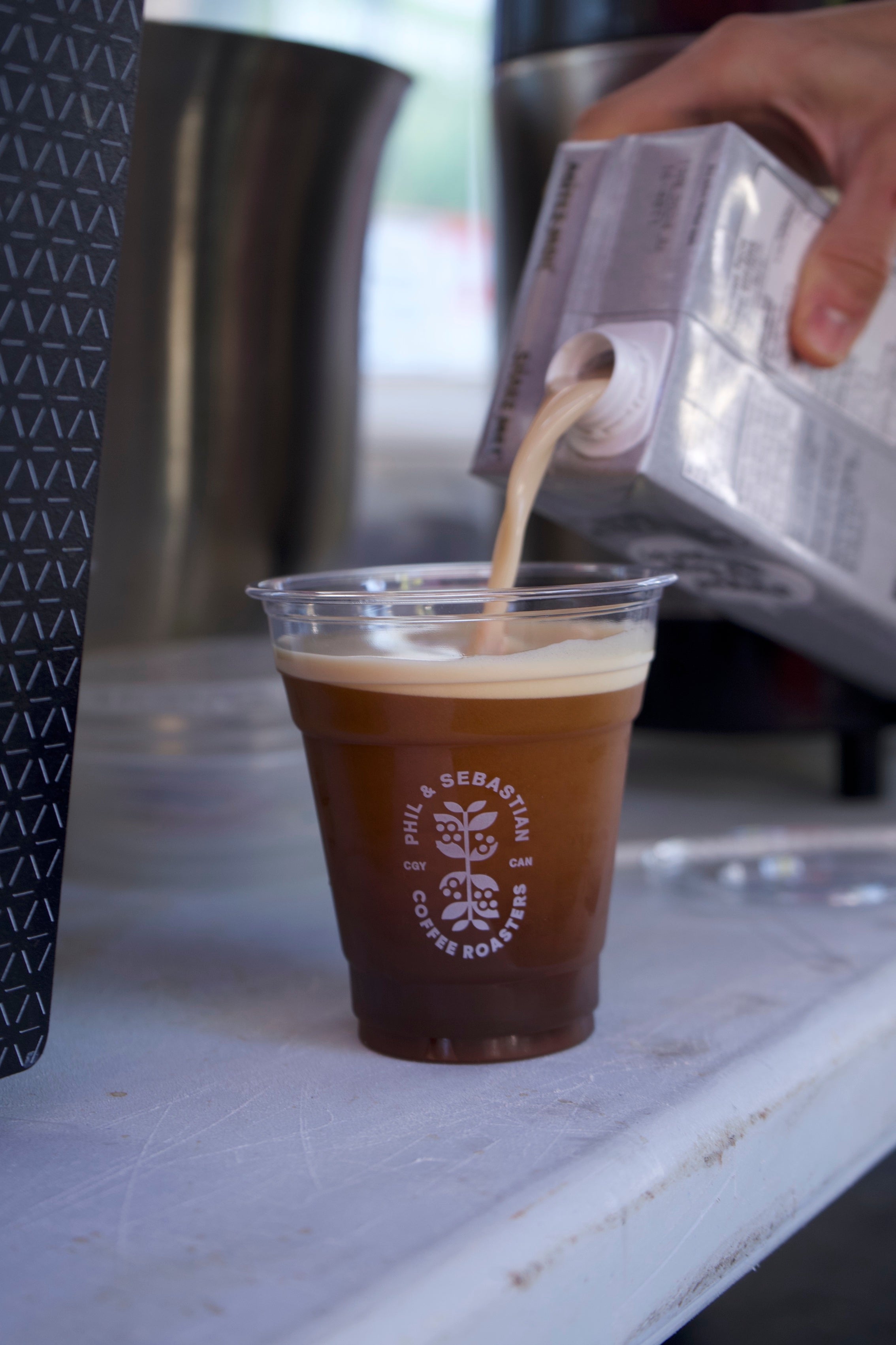 Close up of Oatly being poured into a cup with a Phil and Sebastian logo filled with Nitro Cold Brew next to the BROOD.