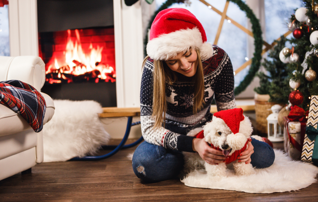 woman and dog sitting on ground by Christmas tree and fireplace wearing Santa hats and Christmas attire together