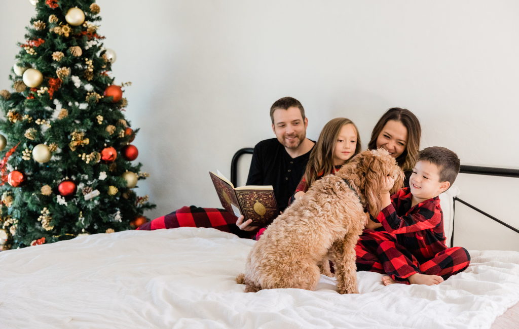 family celebrating Christmas in matching Christmas pajamas next to Christmas tree reading on bed with doodle dog