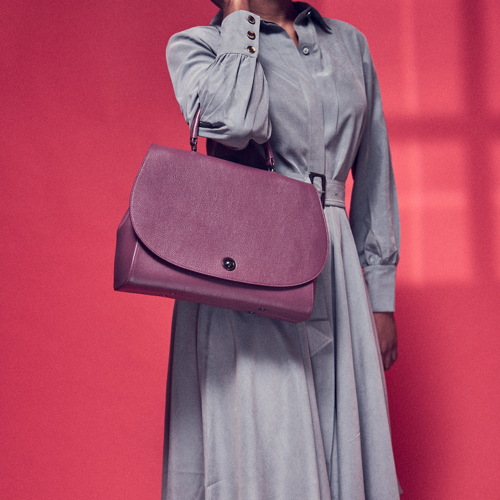 A fashion and style editorial photo showing a woman model holding a burgundy red leather handbag. The bag has a minimalist design with a trapeze shape and rounded oversized flap. This is the Tomoli Briffani Jut interchangeable satchel in Sangria.