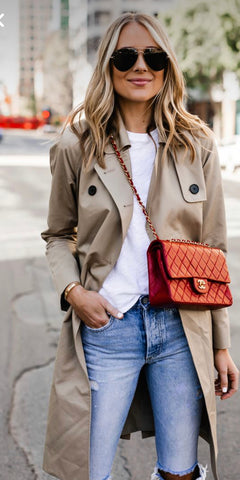 Blonde Caucasian lady in sunglasses wearing a white tshirt under a classic trench coat and with blue denims.  She carries a cross-body Chanel handbag in a rust colour.