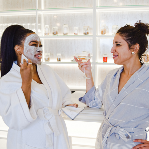 two-women-identifying-individuals-in-robes-and-face-masks-laughing-and-chatting-with martinis-in-their-hands