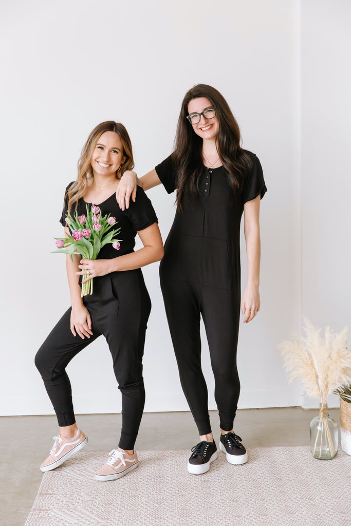 two female models wearing black smash + Tess rompers with bunch of flowers in front of white wall