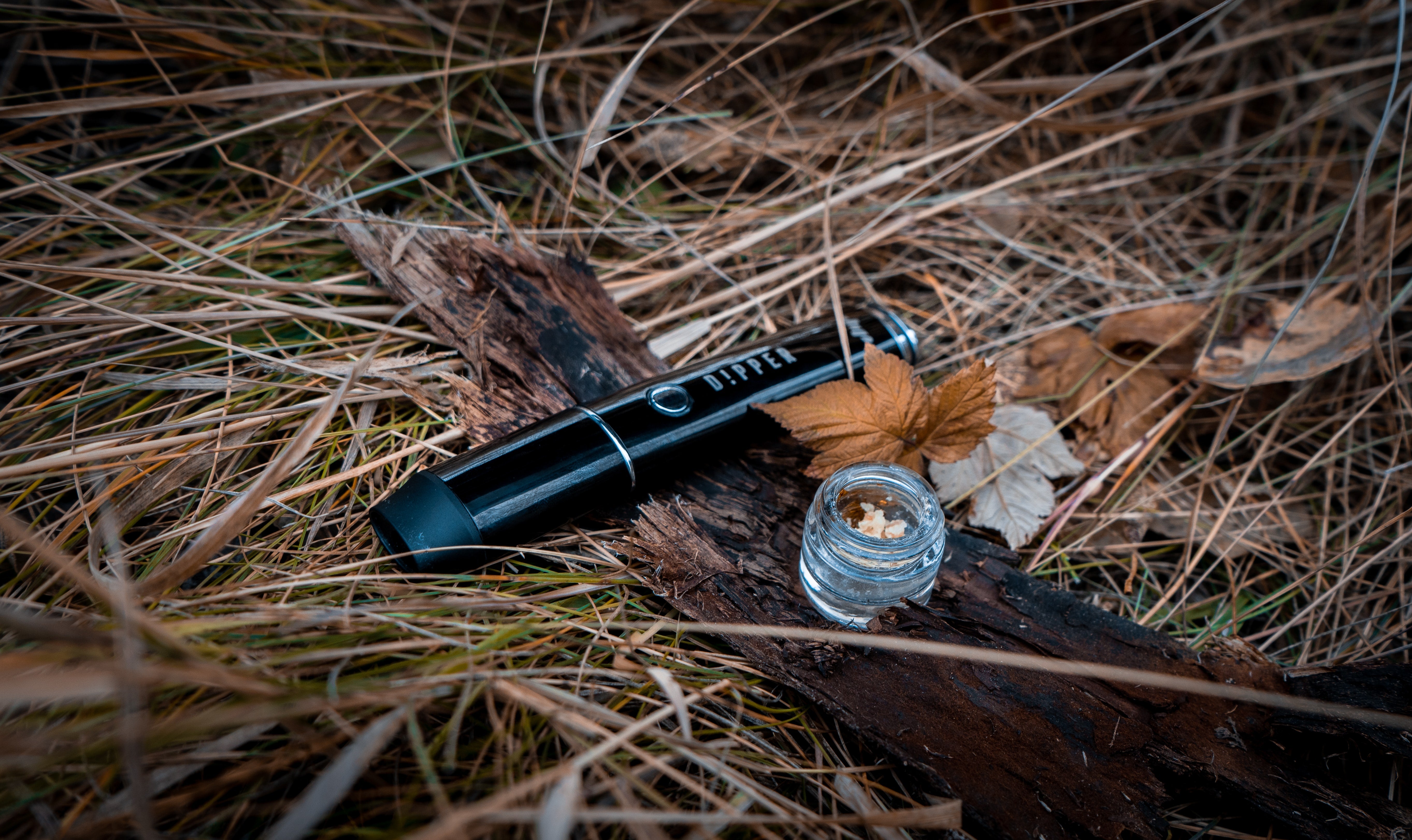 A black dab pen sits next to a glass jar of cannabis extract on wood and grass.