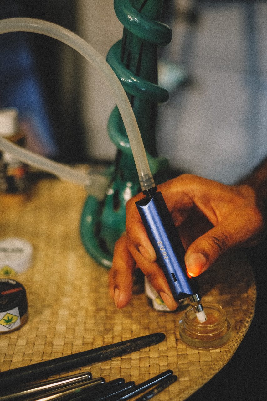 A person dips a blue dab pen into a glass jar with cannabis extracts on a table.