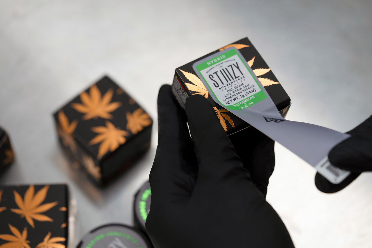 The green label on a small black box with live rosin jam, a cannabis extract, is being removed.