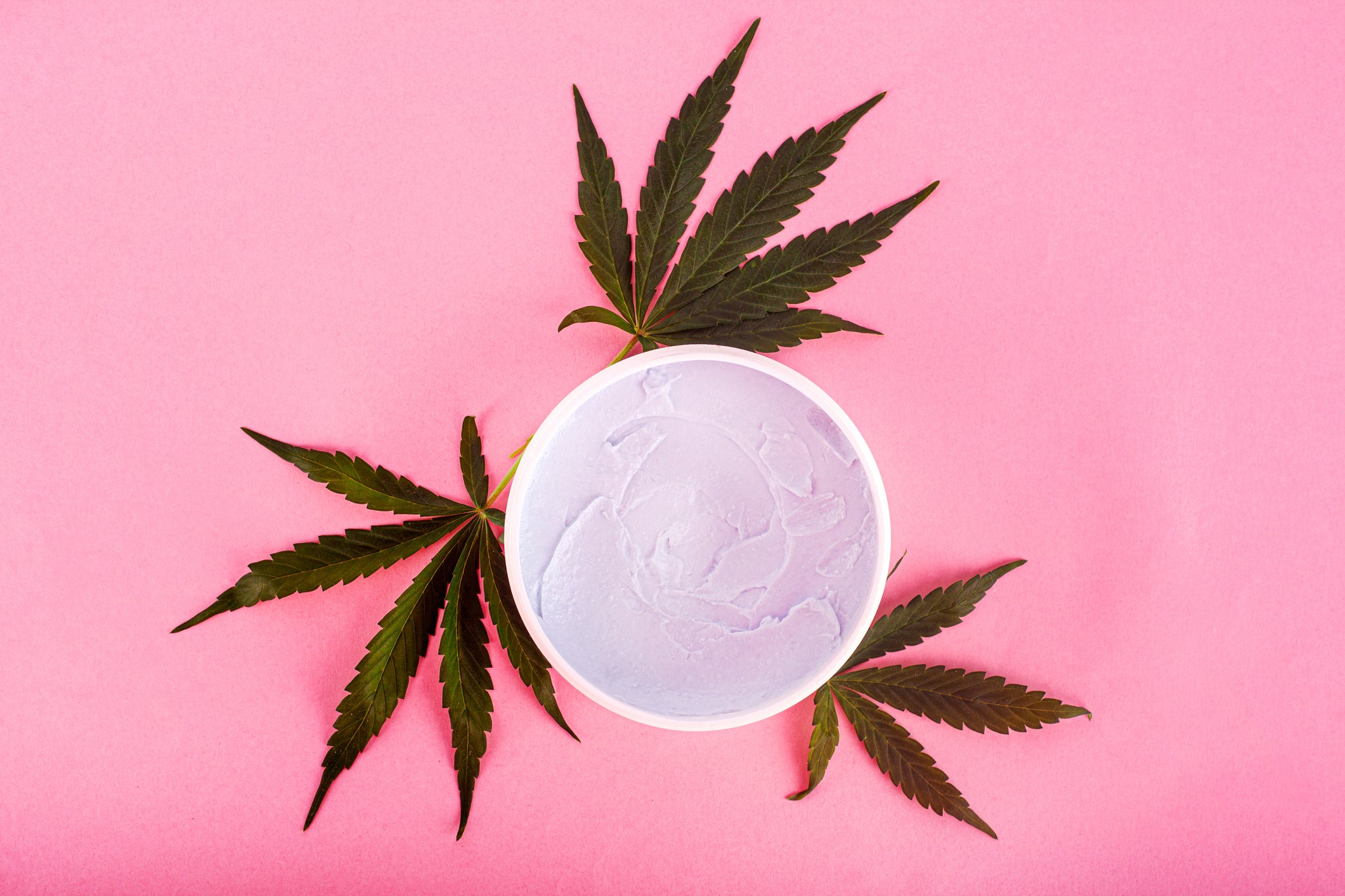 Three cannabis flowers surround a cup of topical cannabis cream on a pink surface.