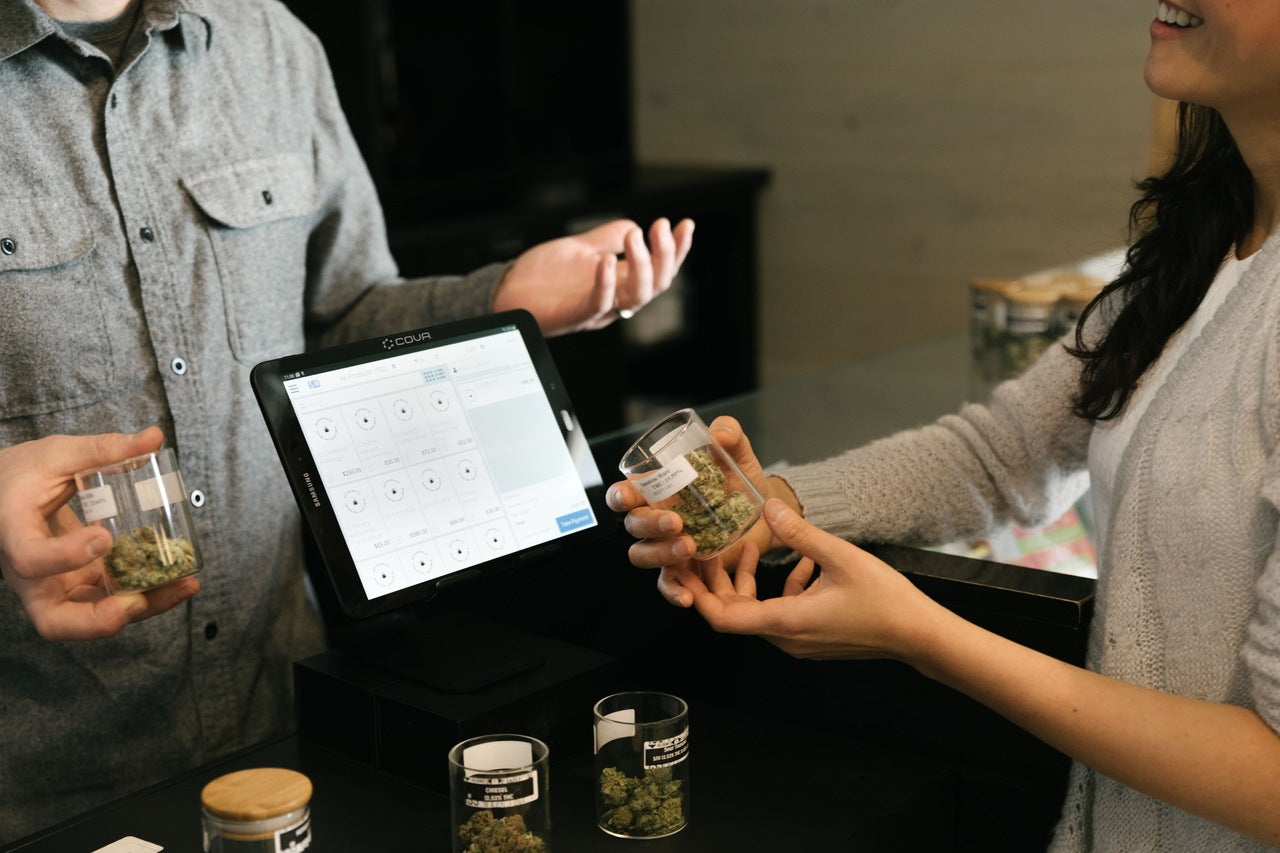 A budtender and a buyer holding glass jars of cannabis flower discuss the different strains of weed.