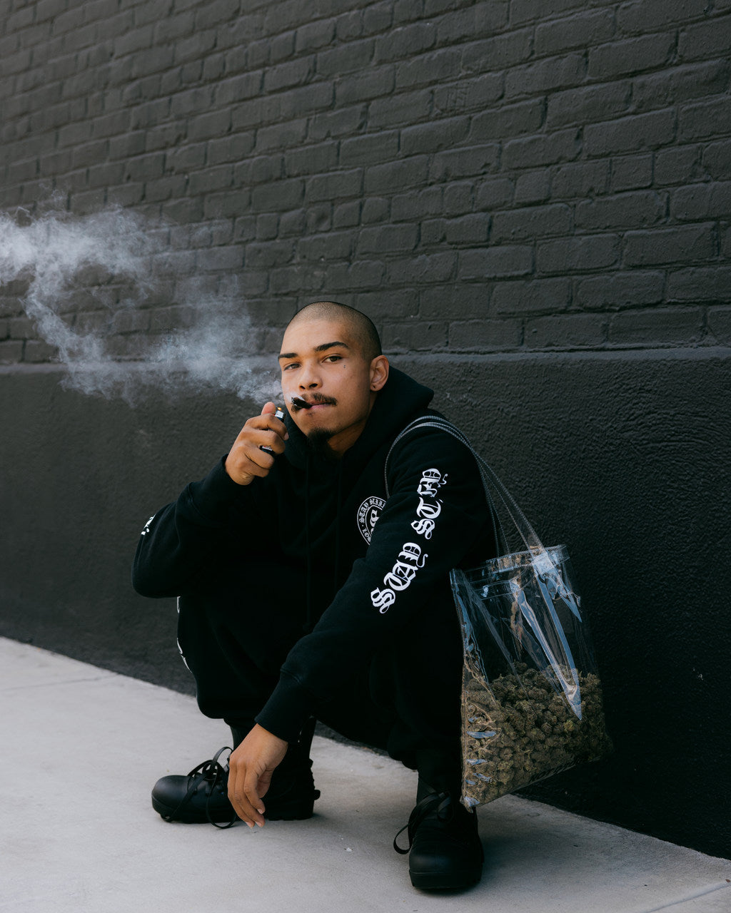 A man dressed in black holds a bag full of cannabis flower while smoking a fat pre-roll.