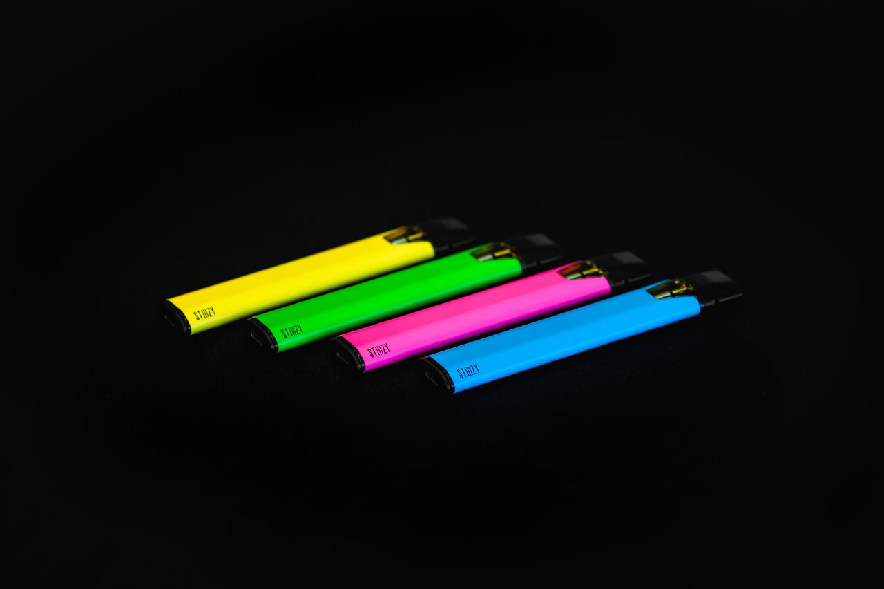 Four neon-colored weed pens with either cannabis distillate or live resin lie on a black surface.