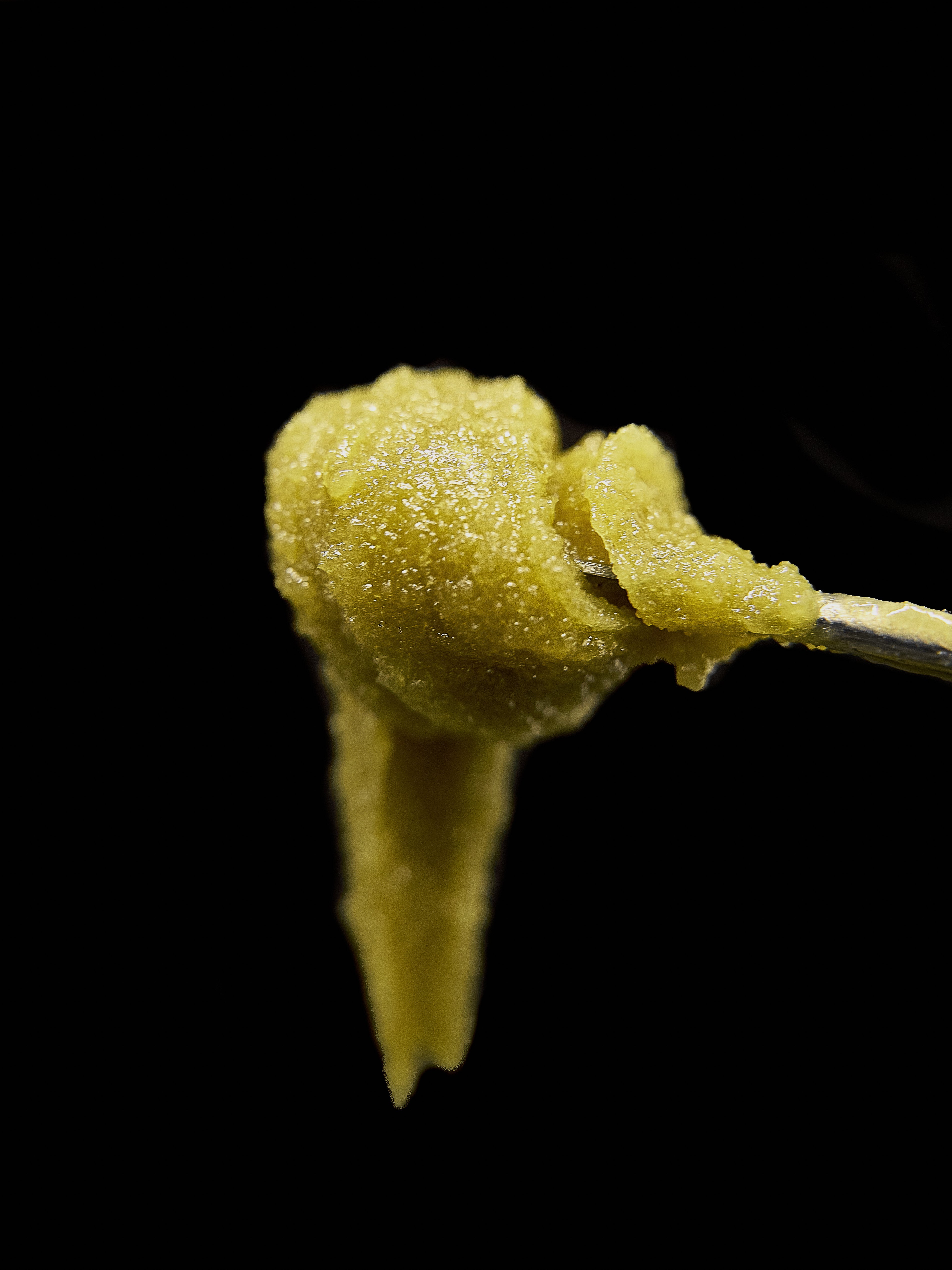 A golden-hued scoop of live rosin cannabis extract is held up with a metal tool against a black background.