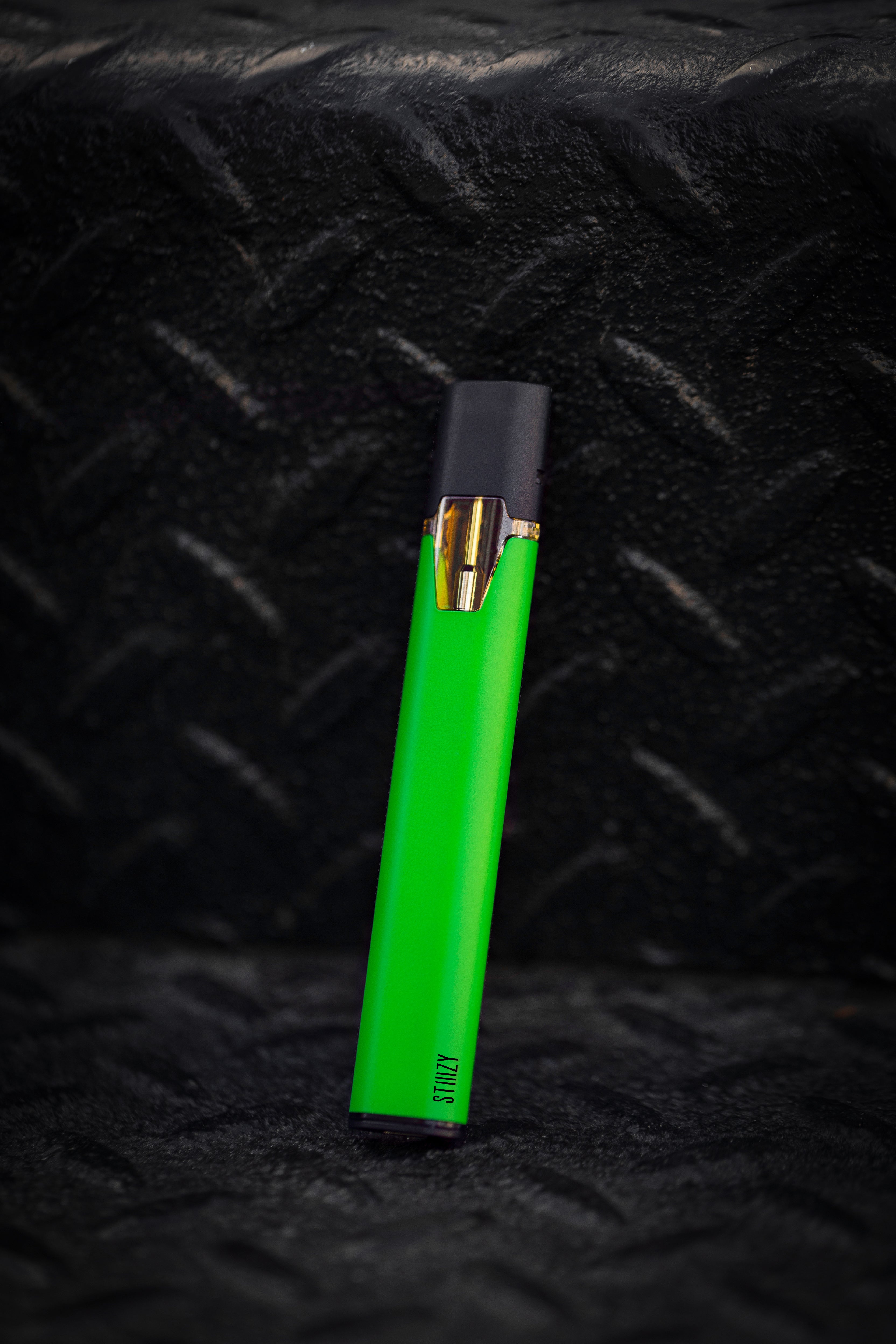 A green STIIIZy weed pen battery and pod for vaping leans against a black wall.
