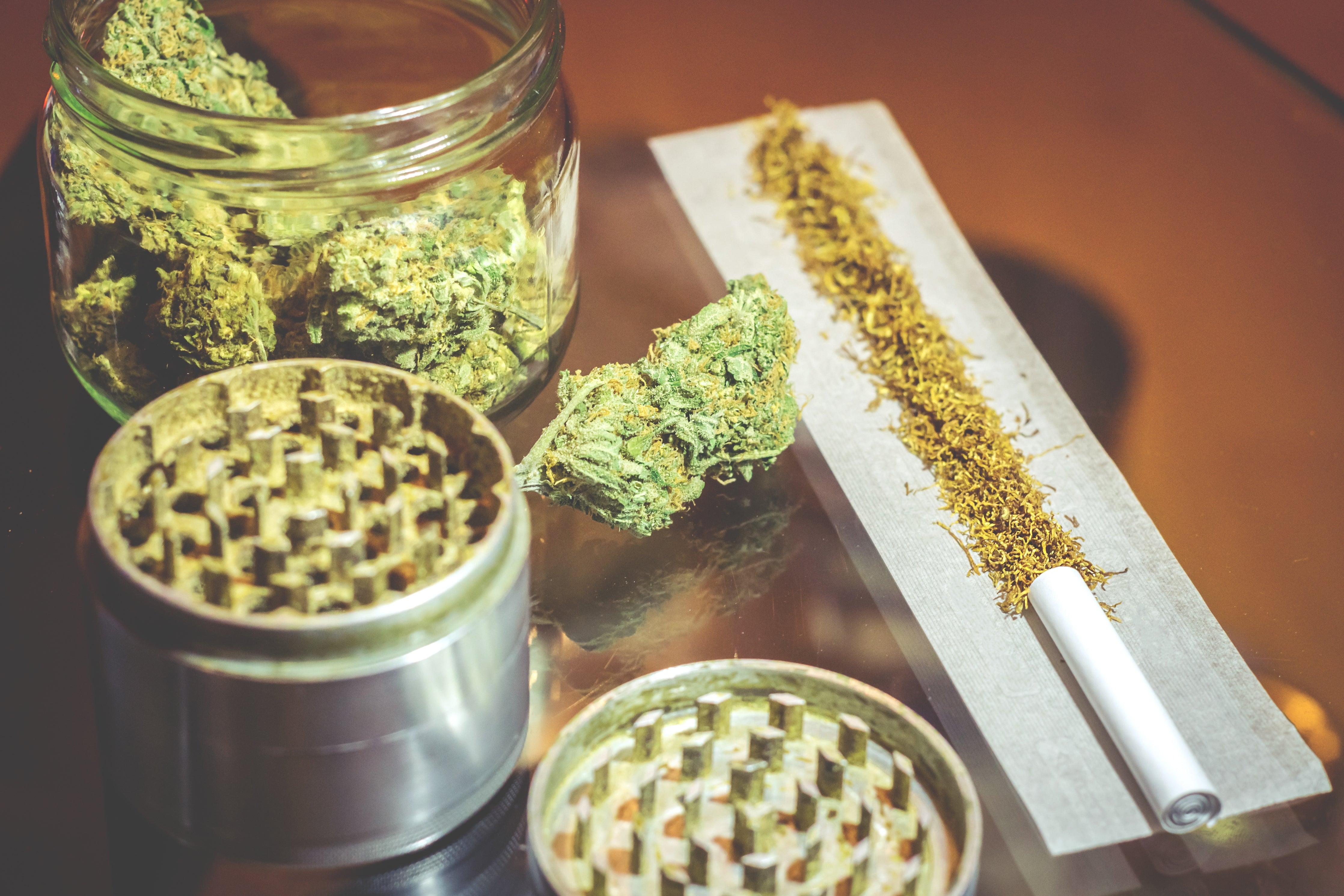 A glass jar of cannabis flower sits next to grinders and ground weed being rolled up in a joint, not a pre-roll.