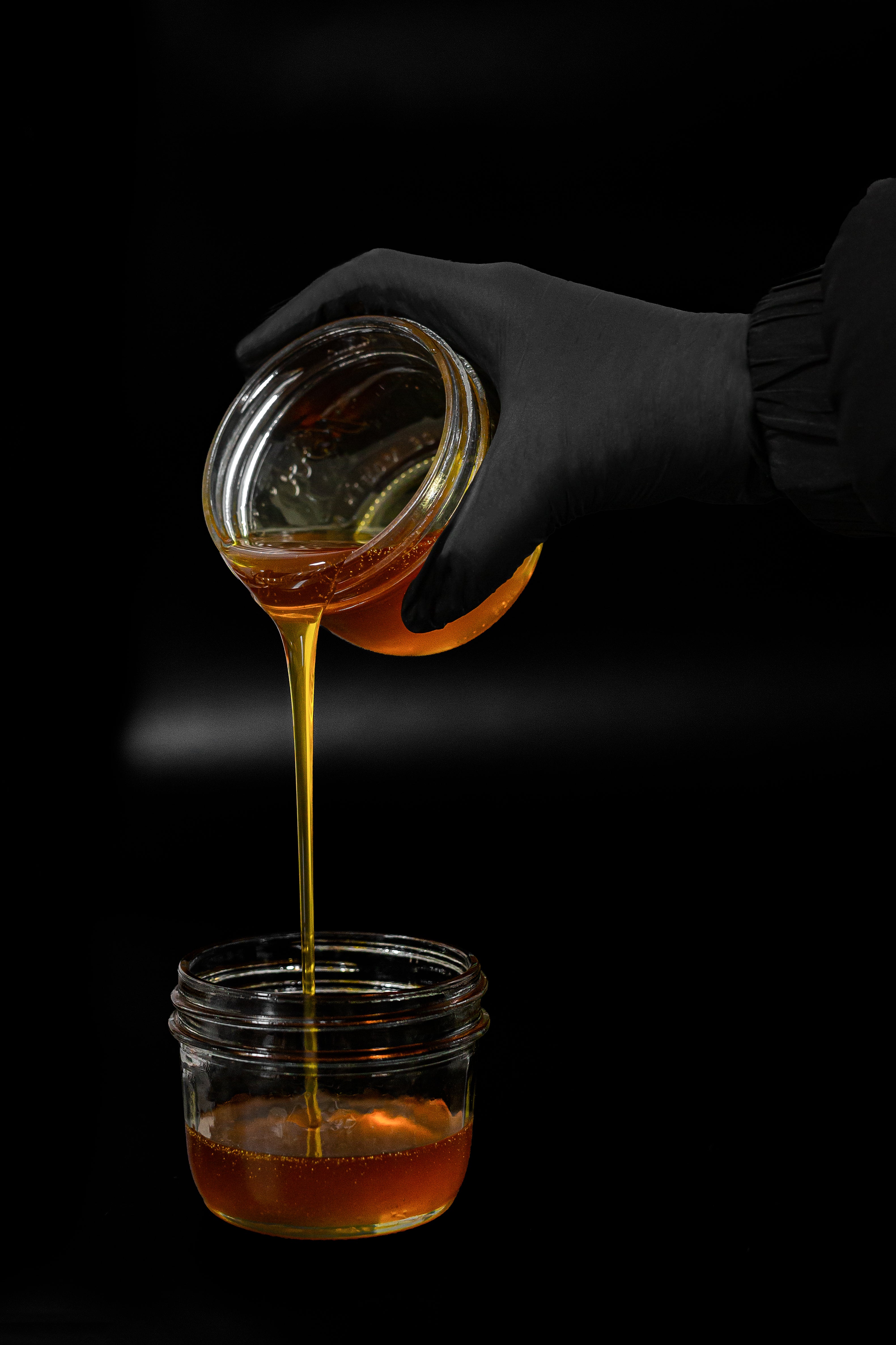 Cannabis distillate for weed vape pods is being poured from one glass jar to another by someone wearing black gloves.