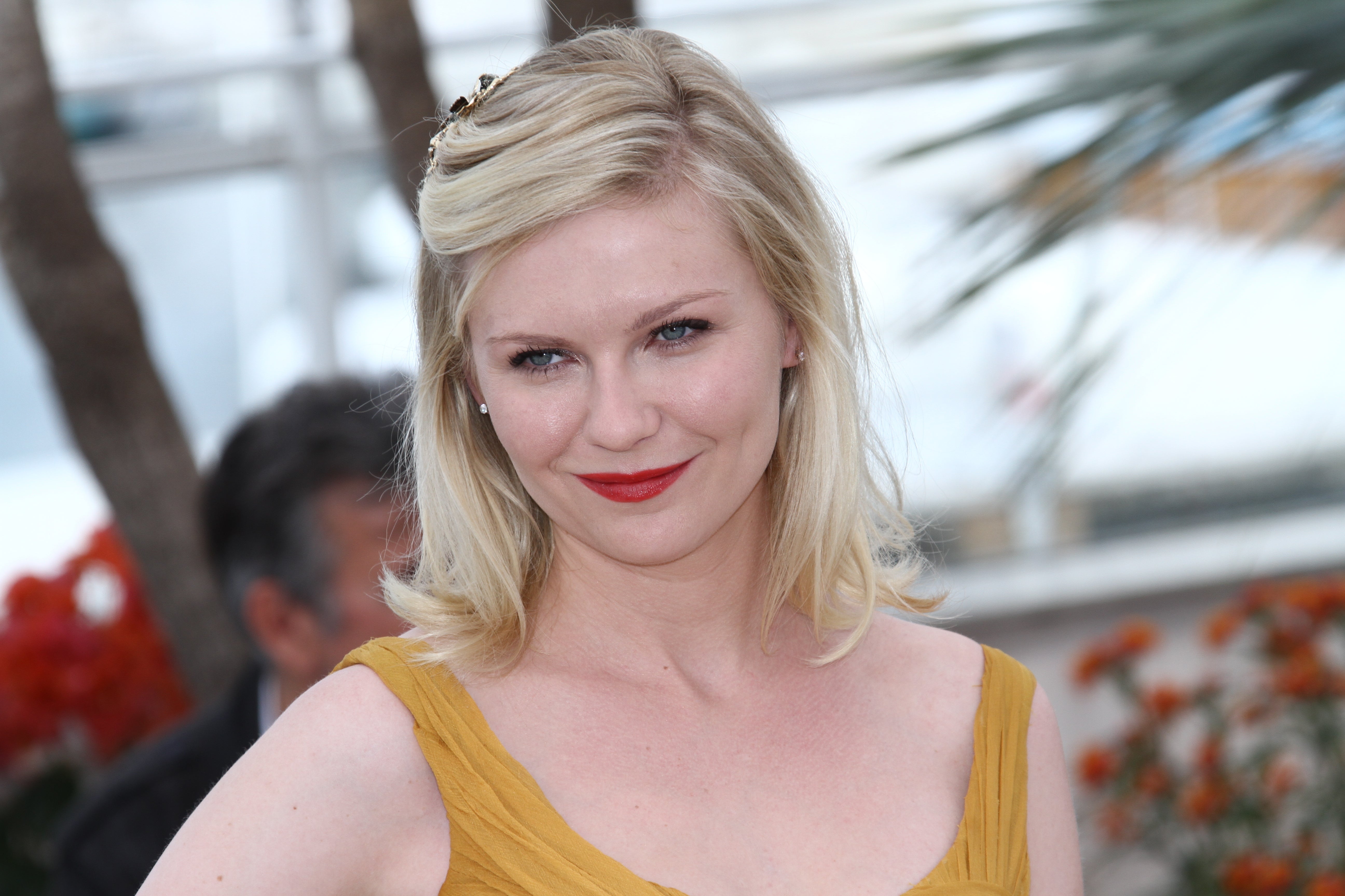 Kirsten Dunst, a cannabis flower-loving celebrity, wears a mustard colored dress and smiles at the camera.