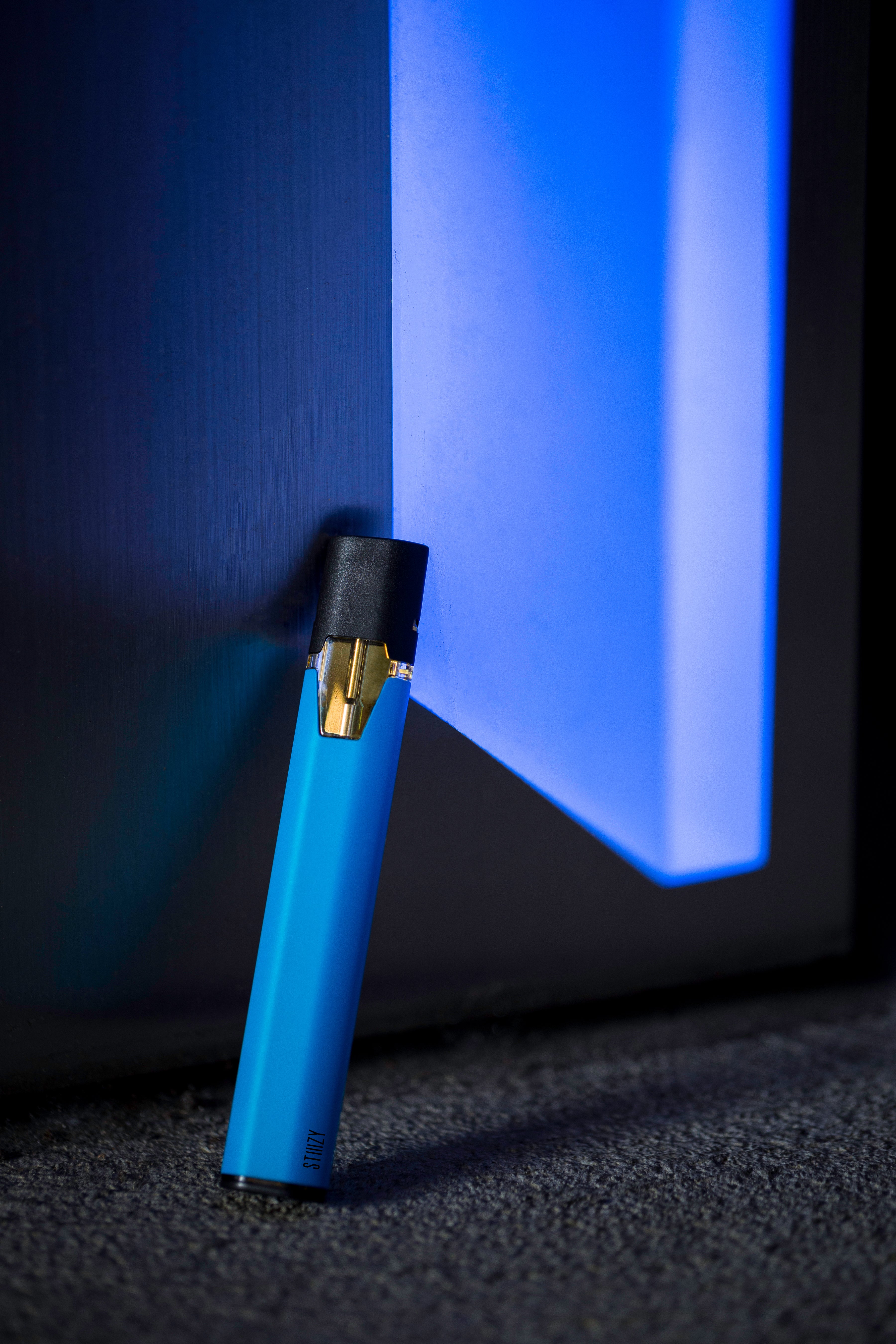 A neon blue STIIIZY weed pen battery with its vape pod attached leans against a neon blue wall.