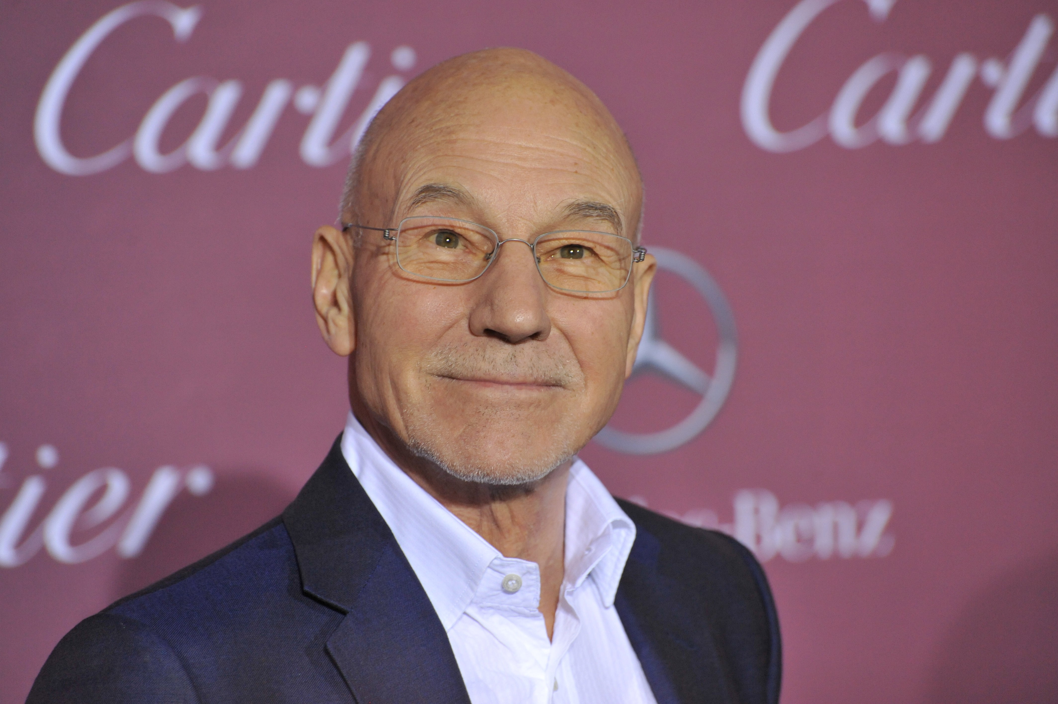 Patrick Stewart, a celebrity who loves cannabis flower, smiles at the camera and wears glasses and a gray suit.