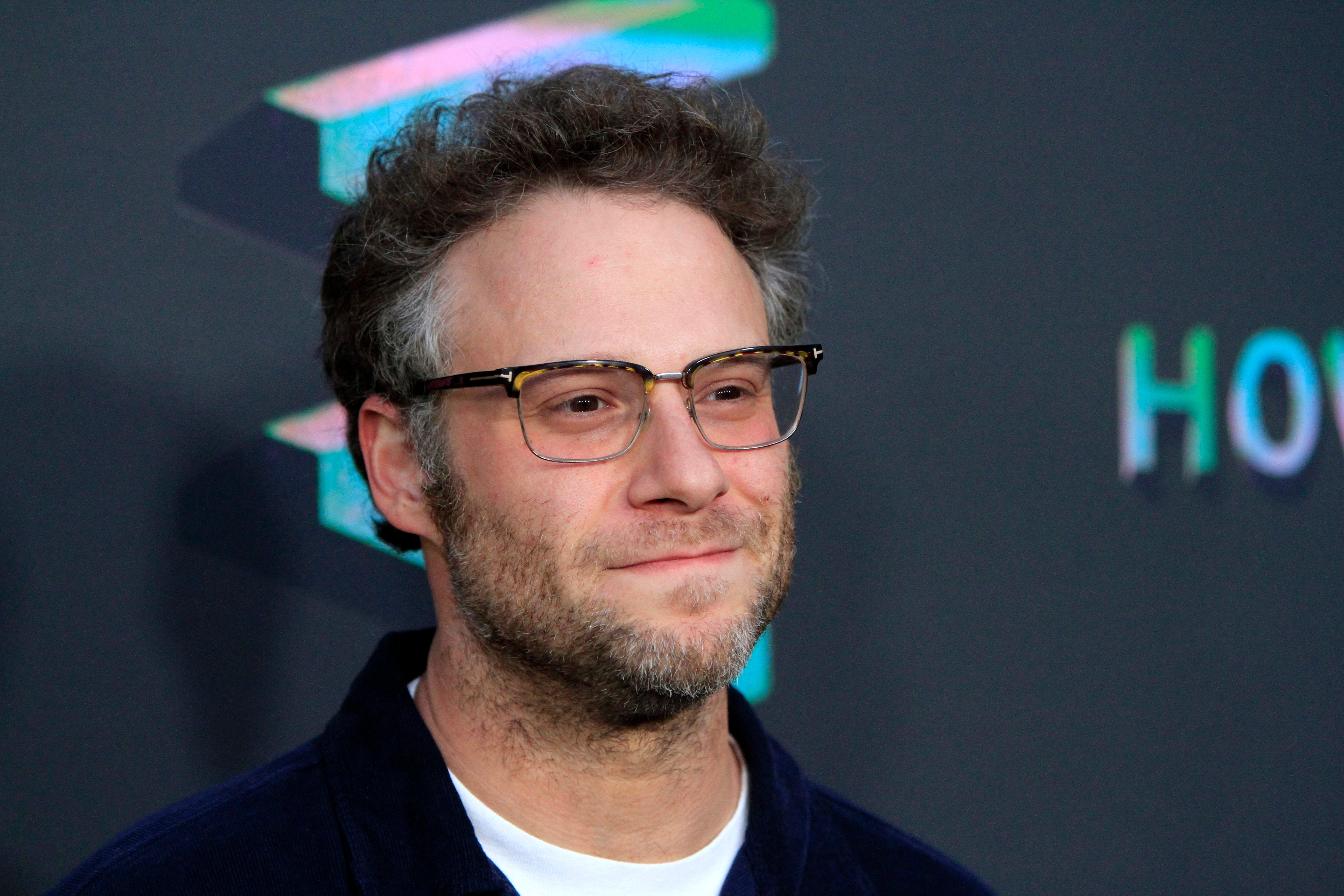 Seth Rogen, a celebrity who loves cannabis flower, is smiling and wearing glasses and a dark-colored collar shirt.