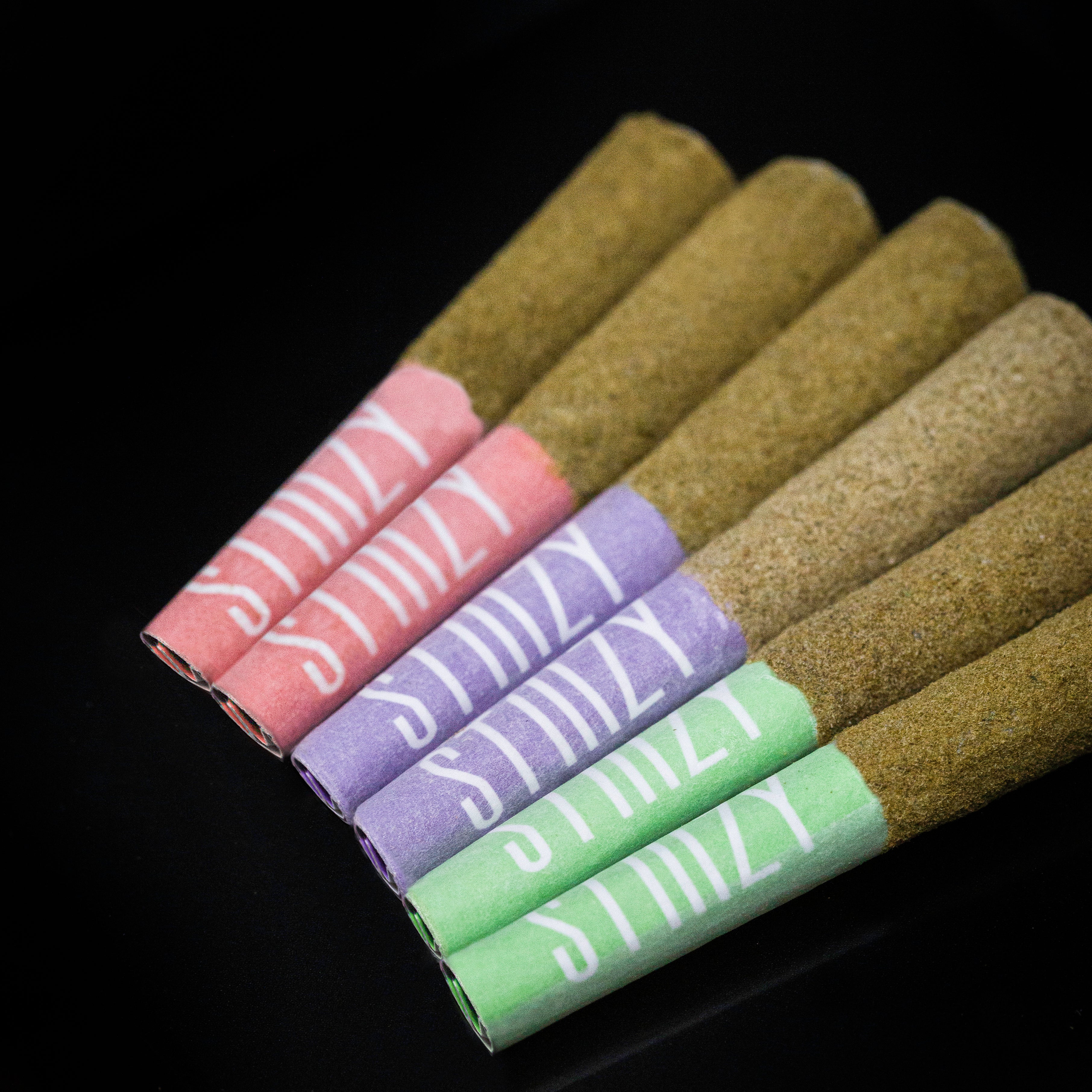 6 STIIIZY infused cannabis pre-rolls with green, purple, and pink filters lie on a black surface.