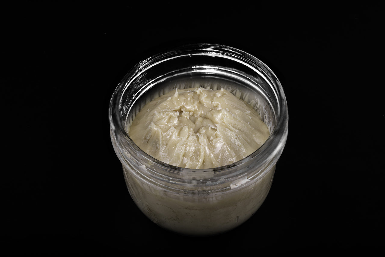 A glass jar in front of a black background contains thick creamy live rosin badder.