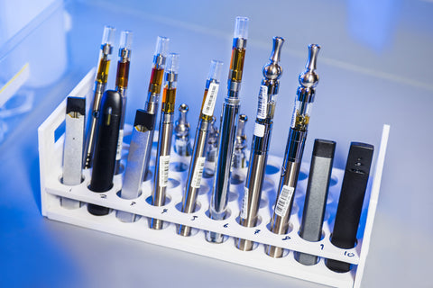Vape pens in various colors and different pods, carts, and batteries are stacked in a rack.