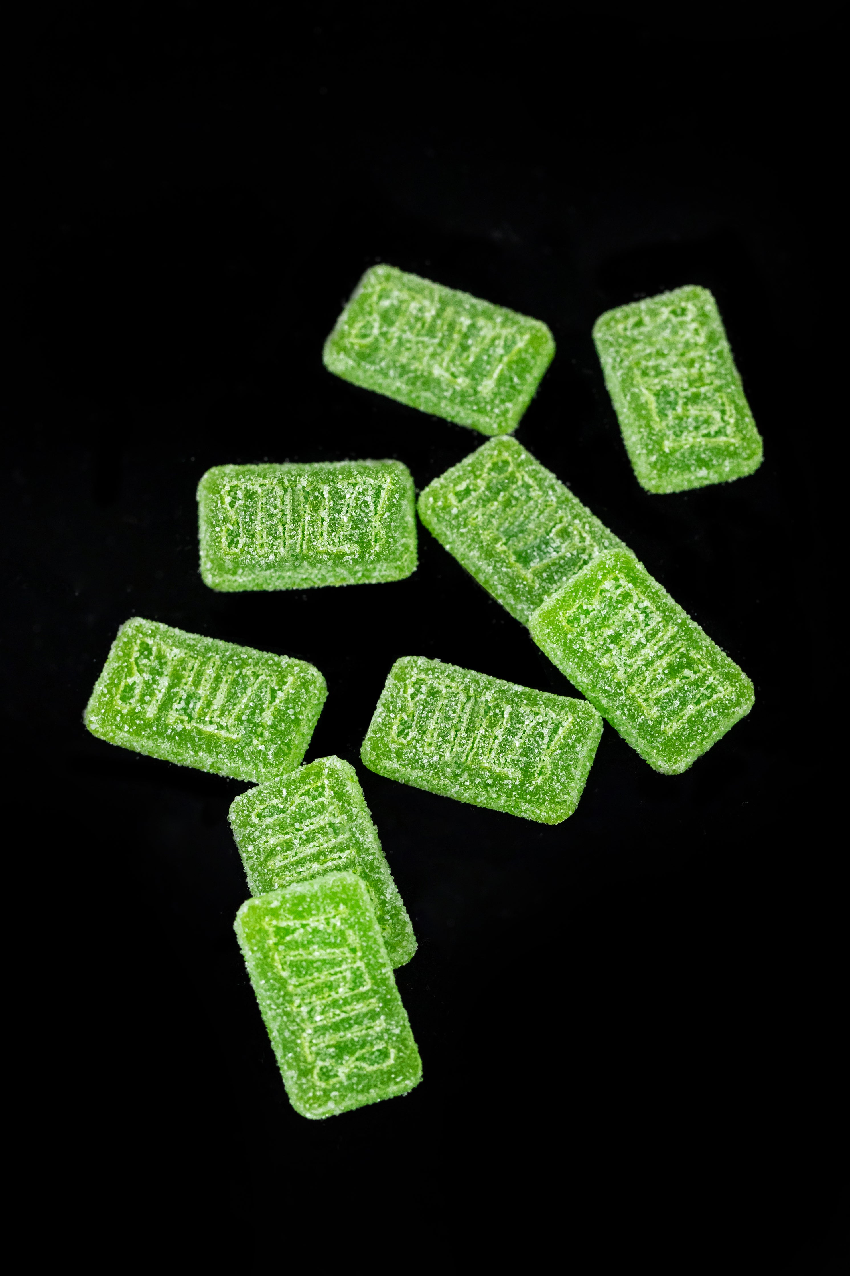 A bunch of bright green weed gummies carefully dosed with THC are scattered over a black background.