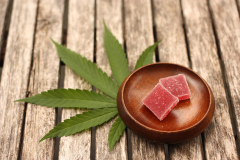 Weed gummies in a wooden bowl like next to a cannabis flower leaf on a wooden table.