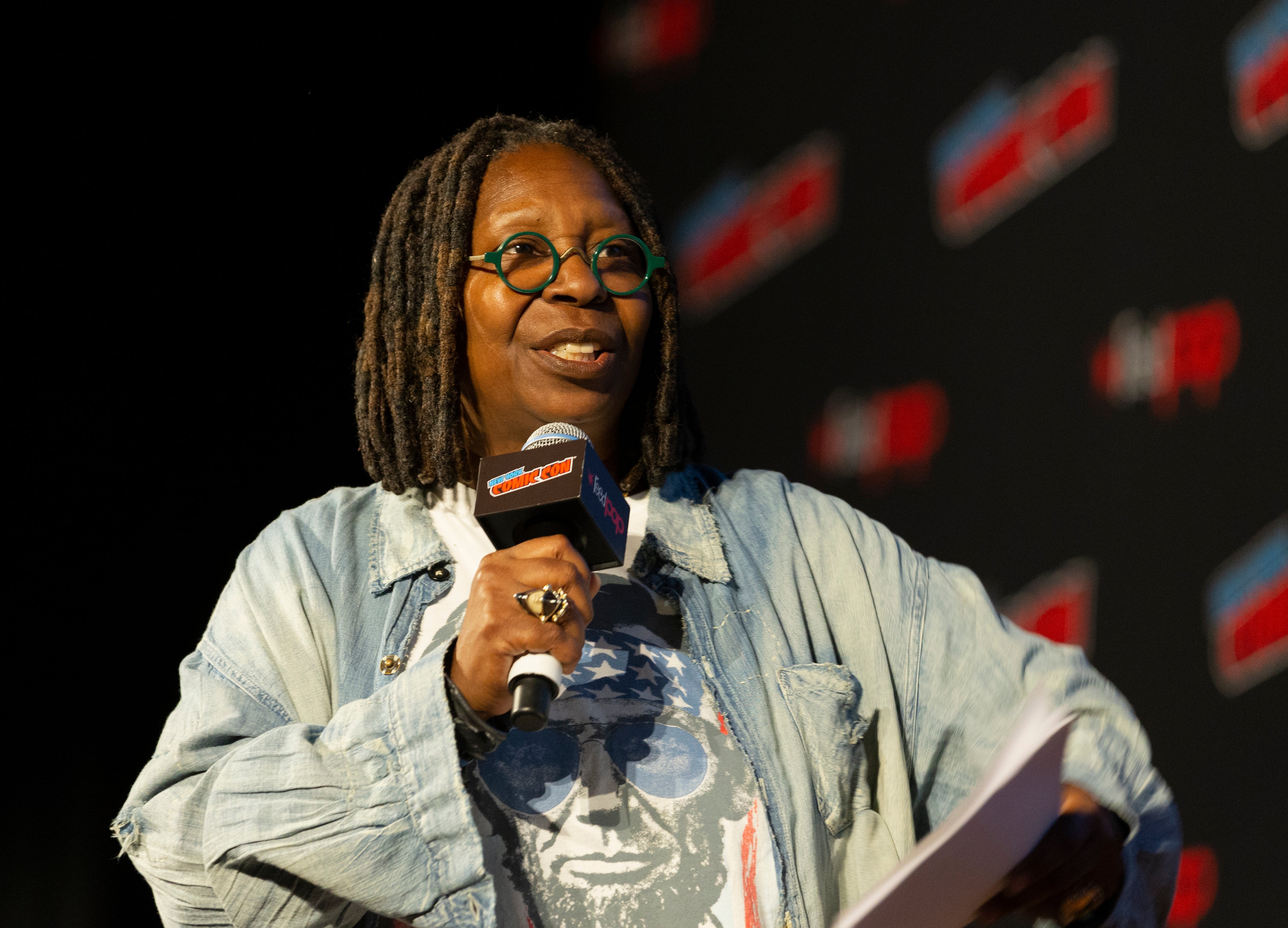 Whoopi Goldberg, a celebrity who loves cannabis flower, talks into a microphone and wears circular green glasses.