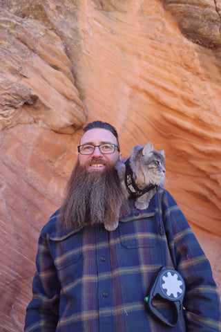Man with beard stands in canyon with cat in harness on shoulder