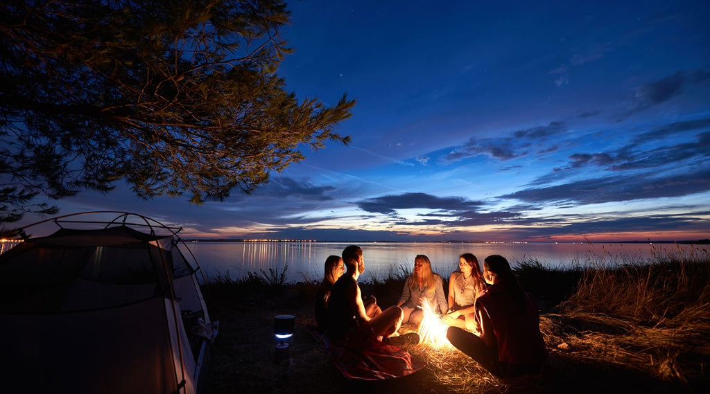 Night summer camping on shore. Group of friends around campfire near tent under evening sky