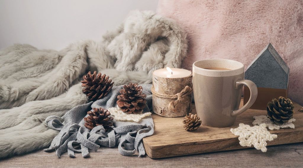 Hygge items including candle cup of coffee pine cones cutting board and cozy throw