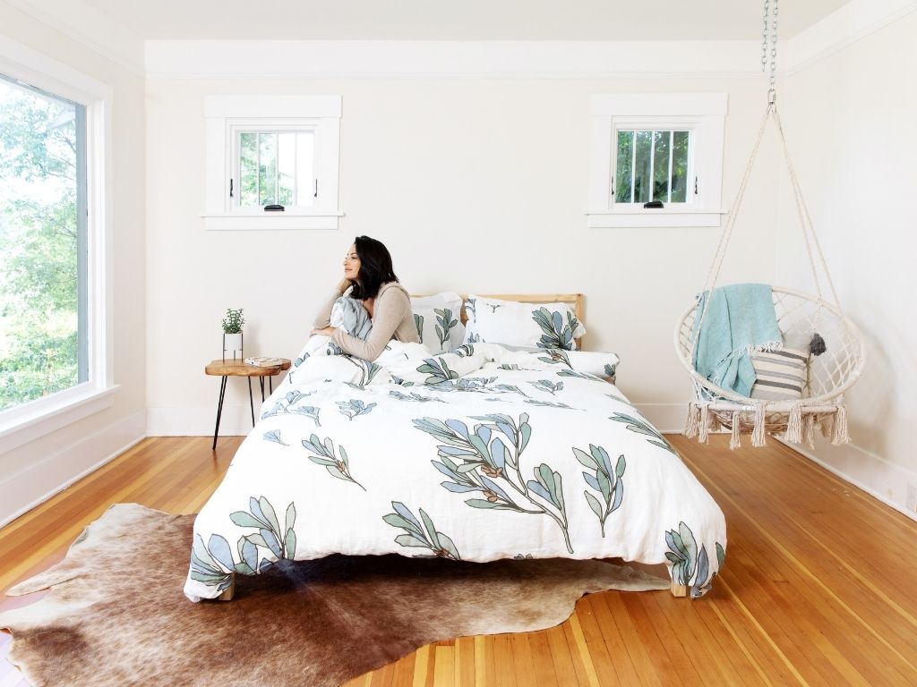 Woman looking out the window sitting on a bed made up with an organic linen duvet cover set with teal and blue leaves by The Modern Dane