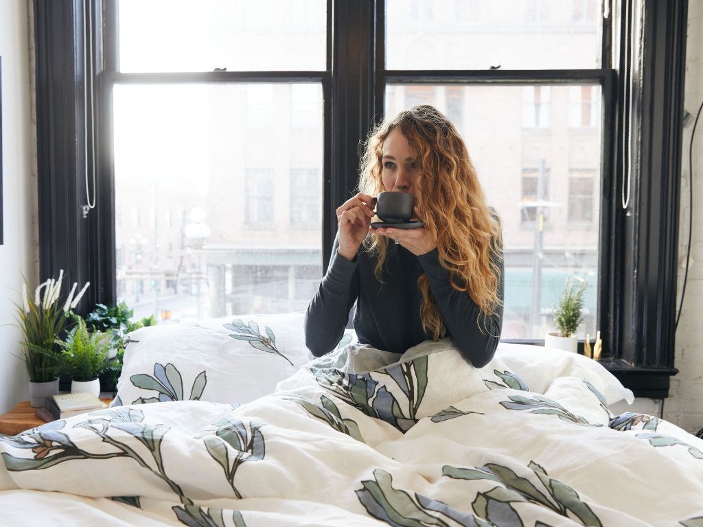 Young woman enjoying a cup of tea in a bed made with organic linen bedding with modern floral print
