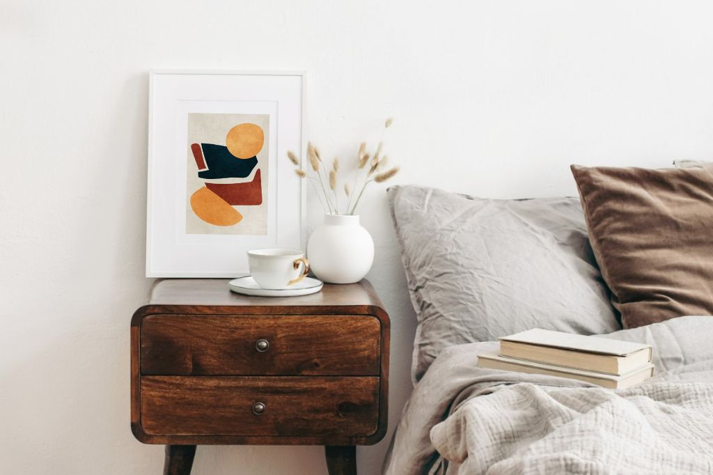 Abstract art on retro wooden bedside table. Modern white ceramic vase with dry Lagurus ovatus grass and cup of coffee. Beige linen and velvet pillows in bedroom. Scandinavian interior.