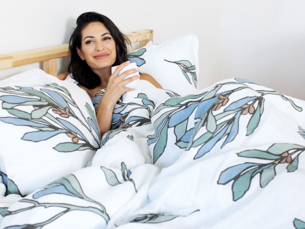 Woman enjoying a cup of tea in organic European linen bedding with teal, green, and blue leaves floral design by The Modern Dane